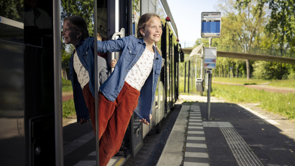 Meisje van een jaar of tien hangt lachend uit stilstaande bus bij een halte in een groen gebied. Meisje van een jaar of tien hangt lachend uit stilstaande bus bij een halte in een groen gebied.