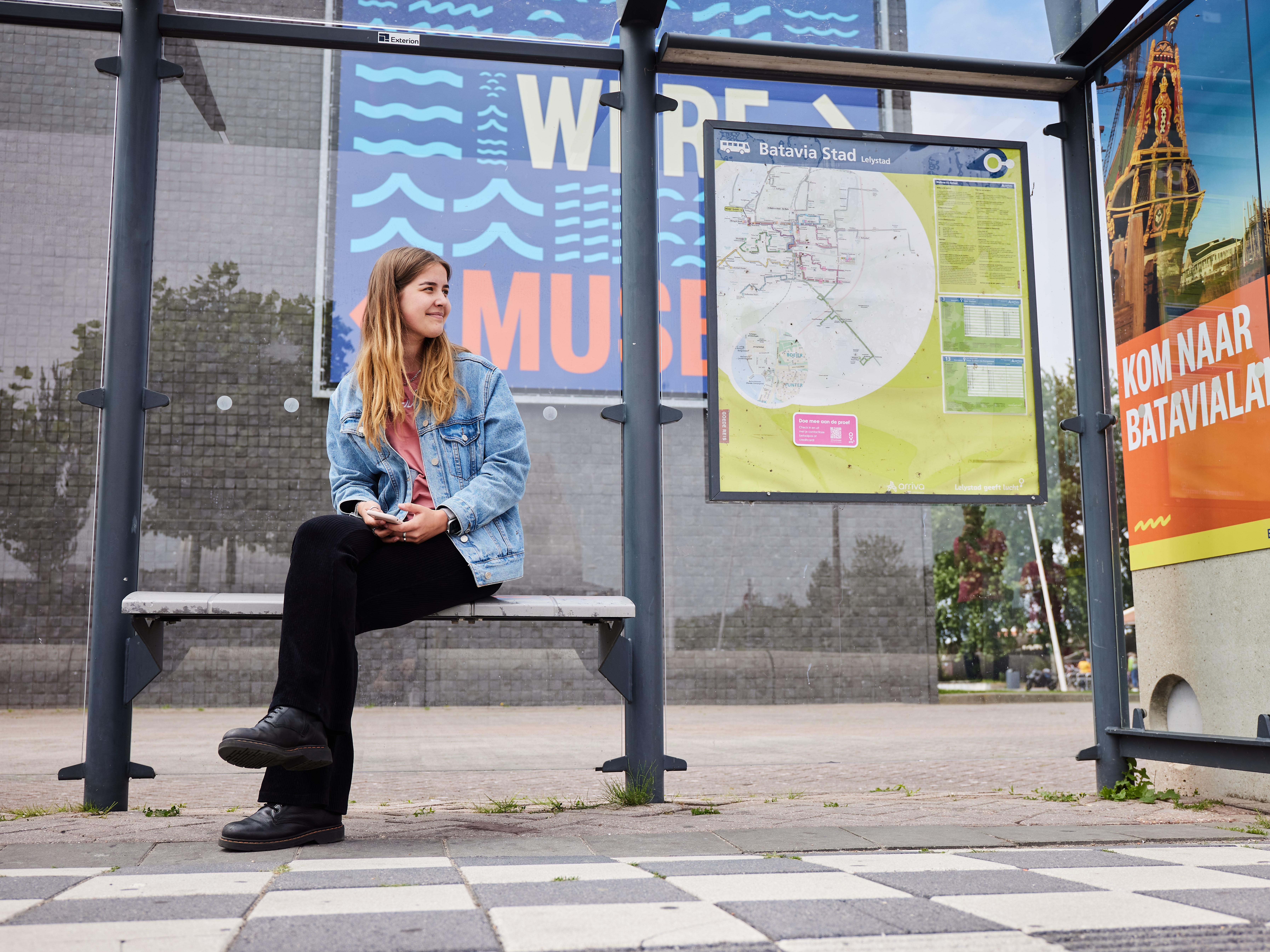 Jonge vrouw zit op bankje van bushalte Batavia Stad met achter haar de vertrekstaat.