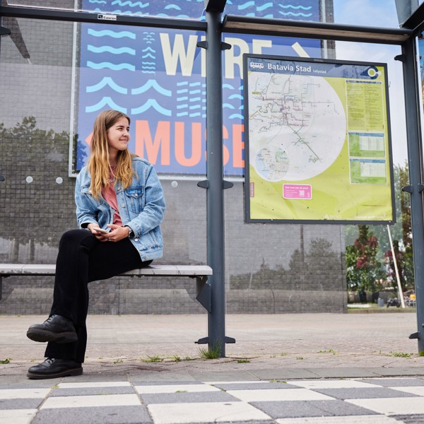 Jonge vrouw zit op bankje van bushalte Batavia Stad met achter haar de vertrekstaat. Jonge vrouw zit op bankje van bushalte Batavia Stad met achter haar de vertrekstaat.