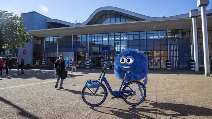 De mascotte van de Arriva-deelfiets, de blauwe fluffy Dirk, staat lachend met zo'n deelfiets voor station Sittard. De mascotte van de Arriva-deelfiets, de blauwe fluffy Dirk, staat lachend met zo'n deelfiets voor station Sittard.