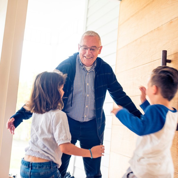 Een oudere man in overhemd en vest lacht en houdt zijn armen wijd voor zijn twee kleinkinderen die aan komen rennen. Een oudere man in overhemd en vest lacht en houdt zijn armen wijd voor zijn twee kleinkinderen die aan komen rennen.