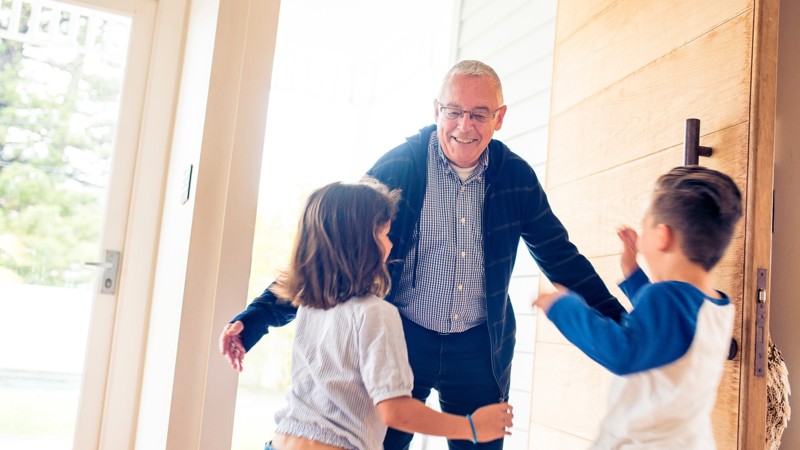 Een oudere man in overhemd en vest lacht en houdt zijn armen wijd voor zijn twee kleinkinderen die aan komen rennen. Een oudere man in overhemd en vest lacht en houdt zijn armen wijd voor zijn twee kleinkinderen die aan komen rennen.