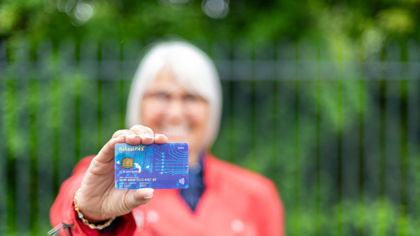 Een oudere vrouw in rode jas houdt een blauwe betaalpas omhoog. Focus ligt op de pas, zij is onscherp op de achtergrond. Een oudere vrouw in rode jas houdt een blauwe betaalpas omhoog. Focus ligt op de pas, zij is onscherp op de achtergrond.