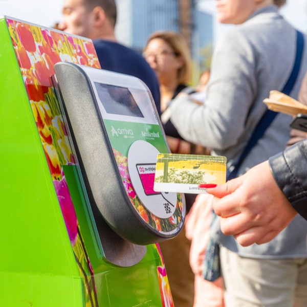 Met kleurige tulpen bestickerde incheckpaal waar een hand een ov-chipkaart bij houdt en met veel mensen in de achtergrond. Met kleurige tulpen bestickerde incheckpaal waar een hand een ov-chipkaart bij houdt en met veel mensen in de achtergrond.