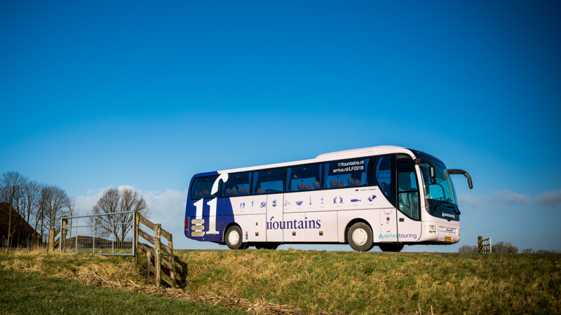Een blauw-witte touringcar van Arriva rijdt met bestickering van '11 fountains' over een Fries dijkje. Een blauw-witte touringcar van Arriva rijdt met bestickering van '11 fountains' over een Fries dijkje.