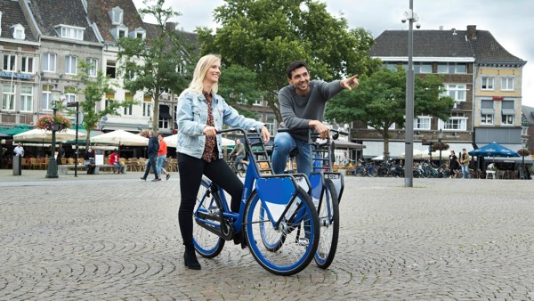 Een man en vrouw staan op een gezellig stadsplein met hun blauwe Arriva-deelfiets, hij wijst en ze lachen allebei. Een man en vrouw staan op een gezellig stadsplein met hun blauwe Arriva-deelfiets, hij wijst en ze lachen allebei.