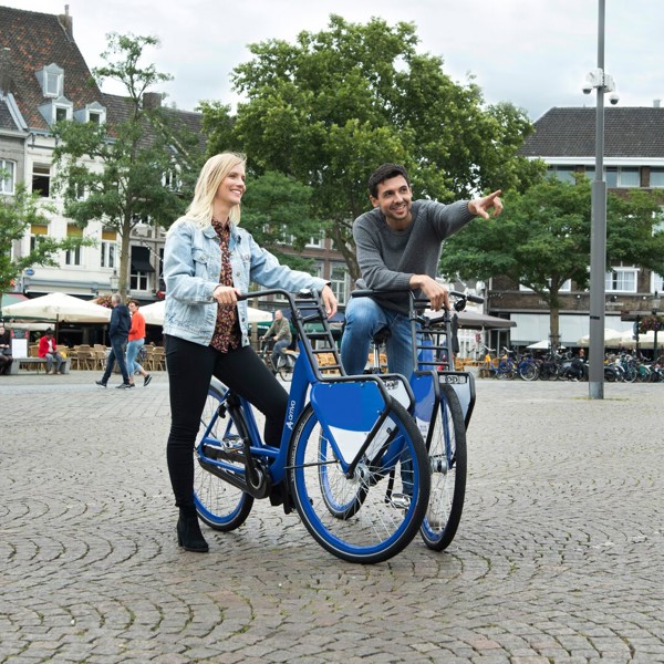 Een man en vrouw staan op een gezellig stadsplein met hun blauwe Arriva-deelfiets, hij wijst en ze lachen allebei. Een man en vrouw staan op een gezellig stadsplein met hun blauwe Arriva-deelfiets, hij wijst en ze lachen allebei.