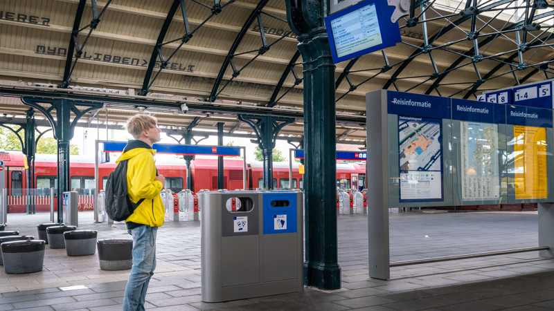 Tiener in felgele jas staat naar de vertrektijden te kijken op een treinperron. Op de achtergrond staat een rode Arriva-trein. Tiener in felgele jas staat naar de vertrektijden te kijken op een treinperron. Op de achtergrond staat een rode Arriva-trein.