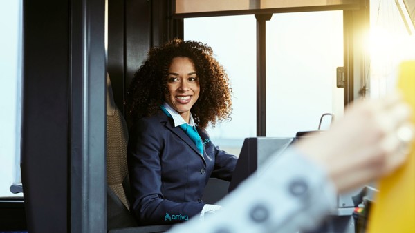 Een vrouwelijke buschauffeur zit achter het stuur en lacht naar iemand die de bus instapt. Een vrouwelijke buschauffeur zit achter het stuur en lacht naar iemand die de bus instapt.