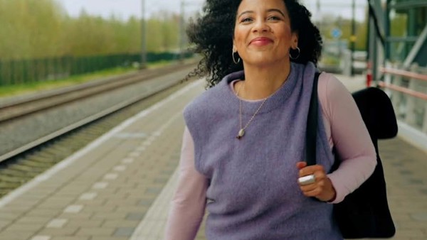 Jonge vrouw staat lachend op een treinperron met een zwarte rugzak duidelijk zichtbaar over haar linkerschouder. Jonge vrouw staat lachend op een treinperron met een zwarte rugzak duidelijk zichtbaar over haar linkerschouder.