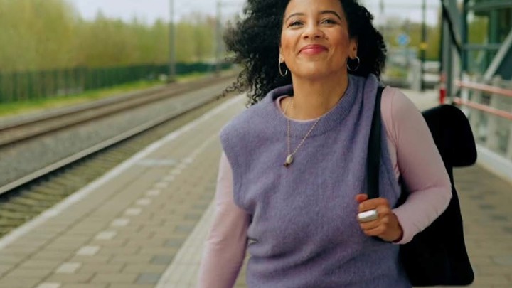 Jonge vrouw staat lachend op een treinperron met een zwarte rugzak duidelijk zichtbaar over haar linkerschouder. Jonge vrouw staat lachend op een treinperron met een zwarte rugzak duidelijk zichtbaar over haar linkerschouder.