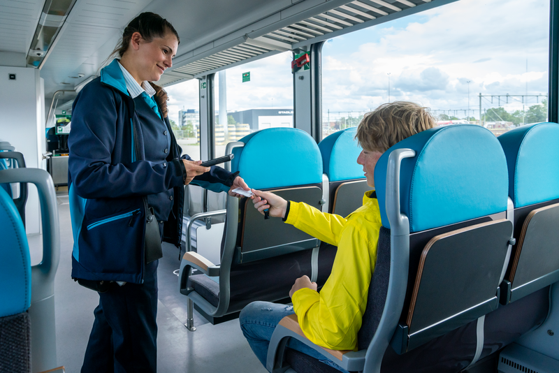 Vrouwelijke steward van Arriva geeft OV-chipkaart terug aan jonge reiziger in gele jas die in de trein zit.