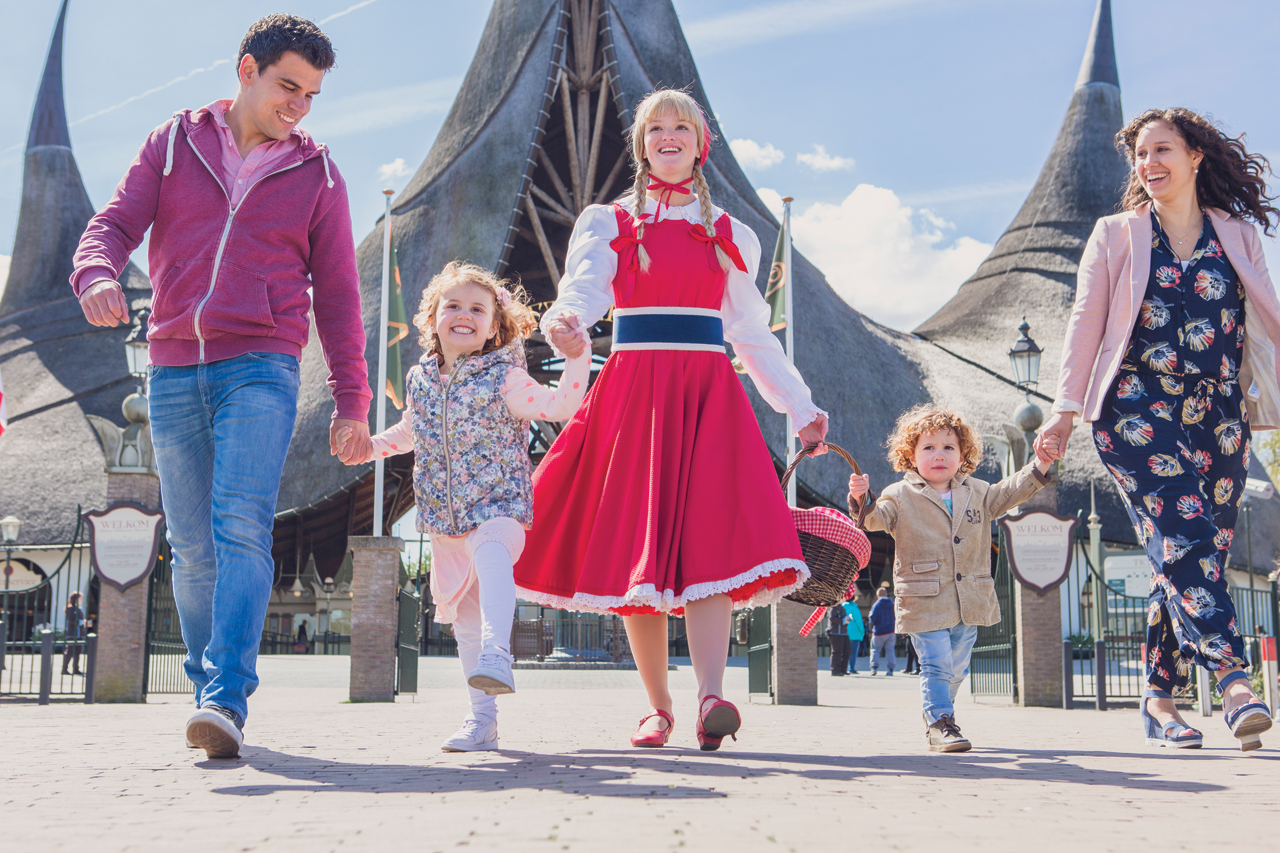 Meisje verkleed als roodkapje loopt met opgetogen jong gezin, hand in hand, de Efteling in, je ziet de ingang achter hen.