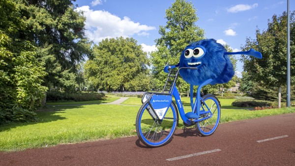 De mascotte van de Arriva-deelfiets, Dirk, zit op zo'n blauwe deelfiets en fiets blij door een stadspark. De mascotte van de Arriva-deelfiets, Dirk, zit op zo'n blauwe deelfiets en fiets blij door een stadspark.