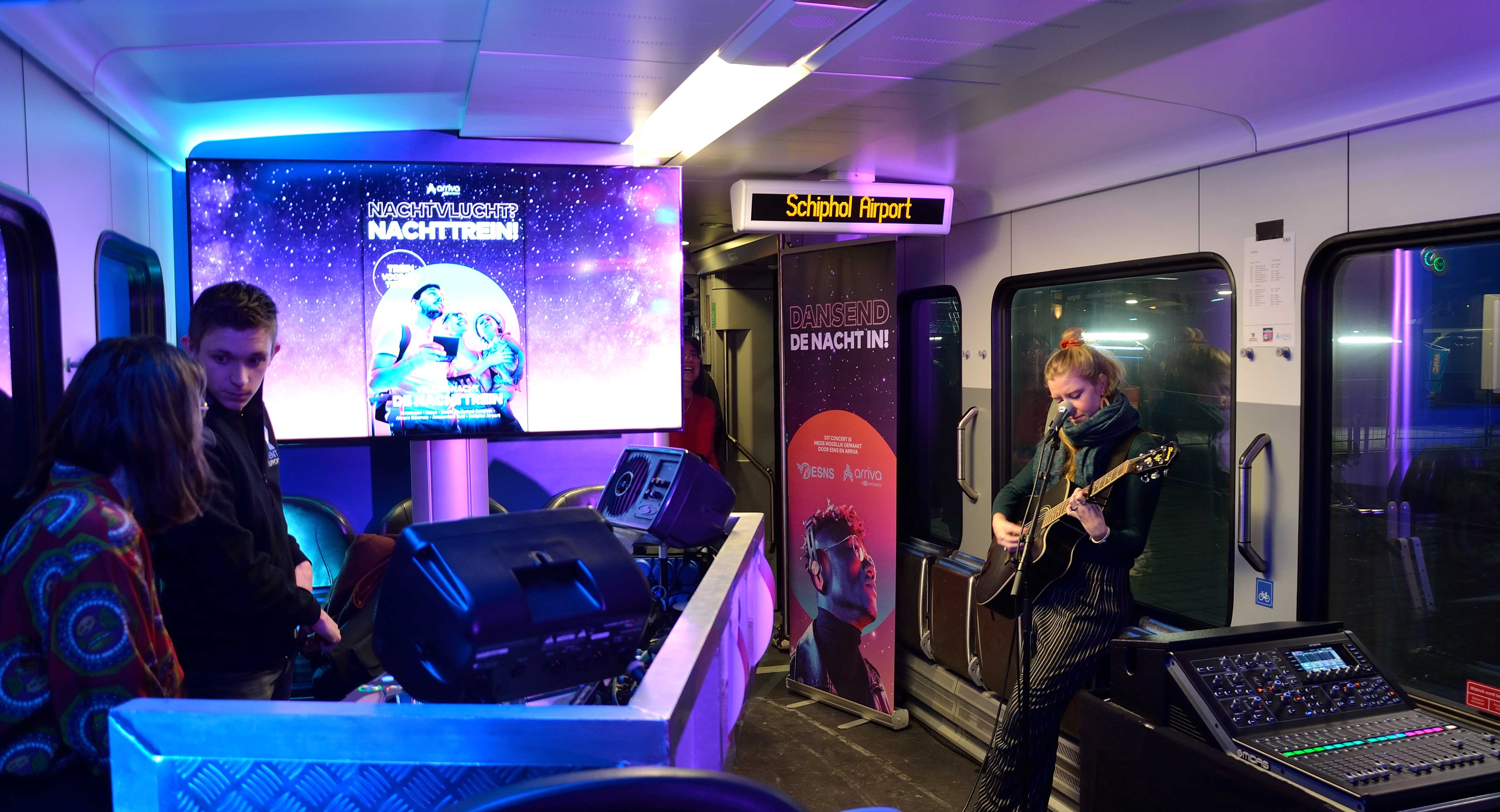 Interieur van een paarsblauw verlichtte nachttrein richting Schiphol met daarin een muzikant met gitaar en twee dj's.