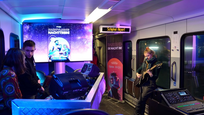 Interieur van een paarsblauw verlichtte nachttrein richting Schiphol met daarin een muzikant met gitaar en twee dj's. Interieur van een paarsblauw verlichtte nachttrein richting Schiphol met daarin een muzikant met gitaar en twee dj's.