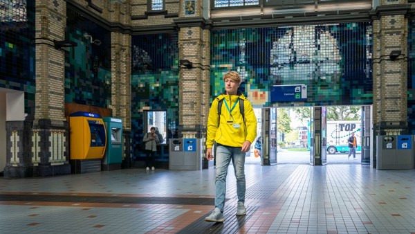 Een jongen in een gele jas loopt door een ruime stationshal met betegelde muren, een klok aan de muur en een hoog plafond. Een jongen in een gele jas loopt door een ruime stationshal met betegelde muren, een klok aan de muur en een hoog plafond.