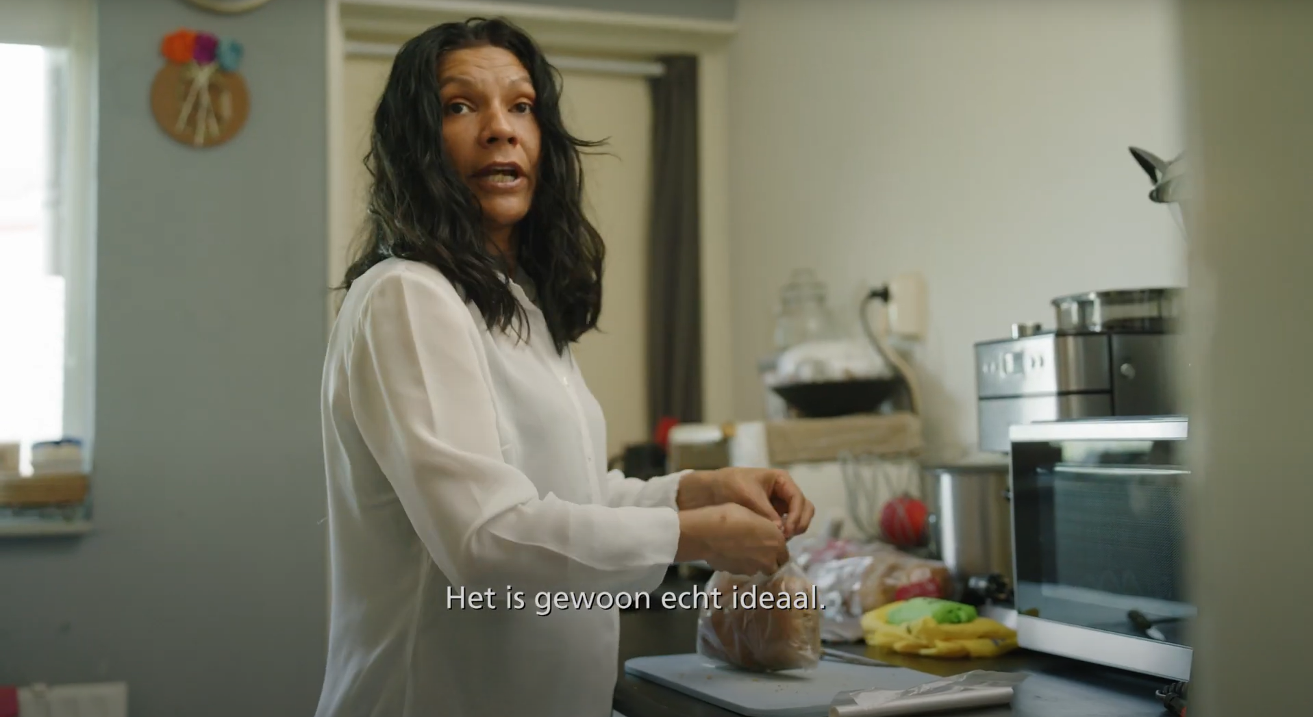 Vrouw staat in keuken een zak met brood open te maken terwijl ze in de camera kijkt en praat.