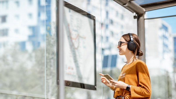 Een lachende jonge vrouw met zonnebril op staat te kijken naar een vertrekstaat in een bushalte. Een lachende jonge vrouw met zonnebril op staat te kijken naar een vertrekstaat in een bushalte.