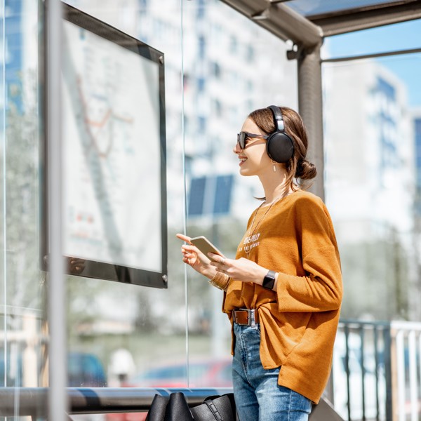 Een lachende jonge vrouw met zonnebril op staat te kijken naar een vertrekstaat in een bushalte. Een lachende jonge vrouw met zonnebril op staat te kijken naar een vertrekstaat in een bushalte.