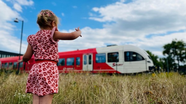 Een meisje van een jaar of vijf in roze zomerjurk staat in hoog gras en kijkt naar een rode Arriva-trein. Een meisje van een jaar of vijf in roze zomerjurk staat in hoog gras en kijkt naar een rode Arriva-trein.