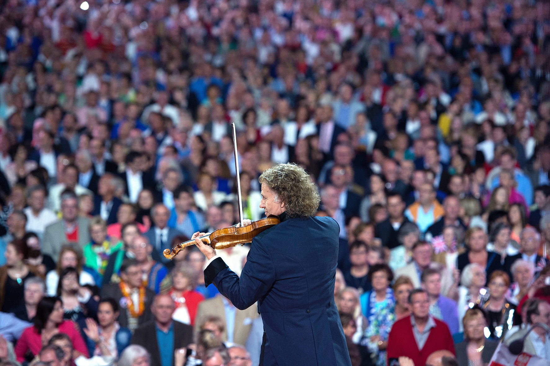 Andre Rieu vanaf de rug gezien speelt in pak viool voor honderden toeschouwers op een tribune.