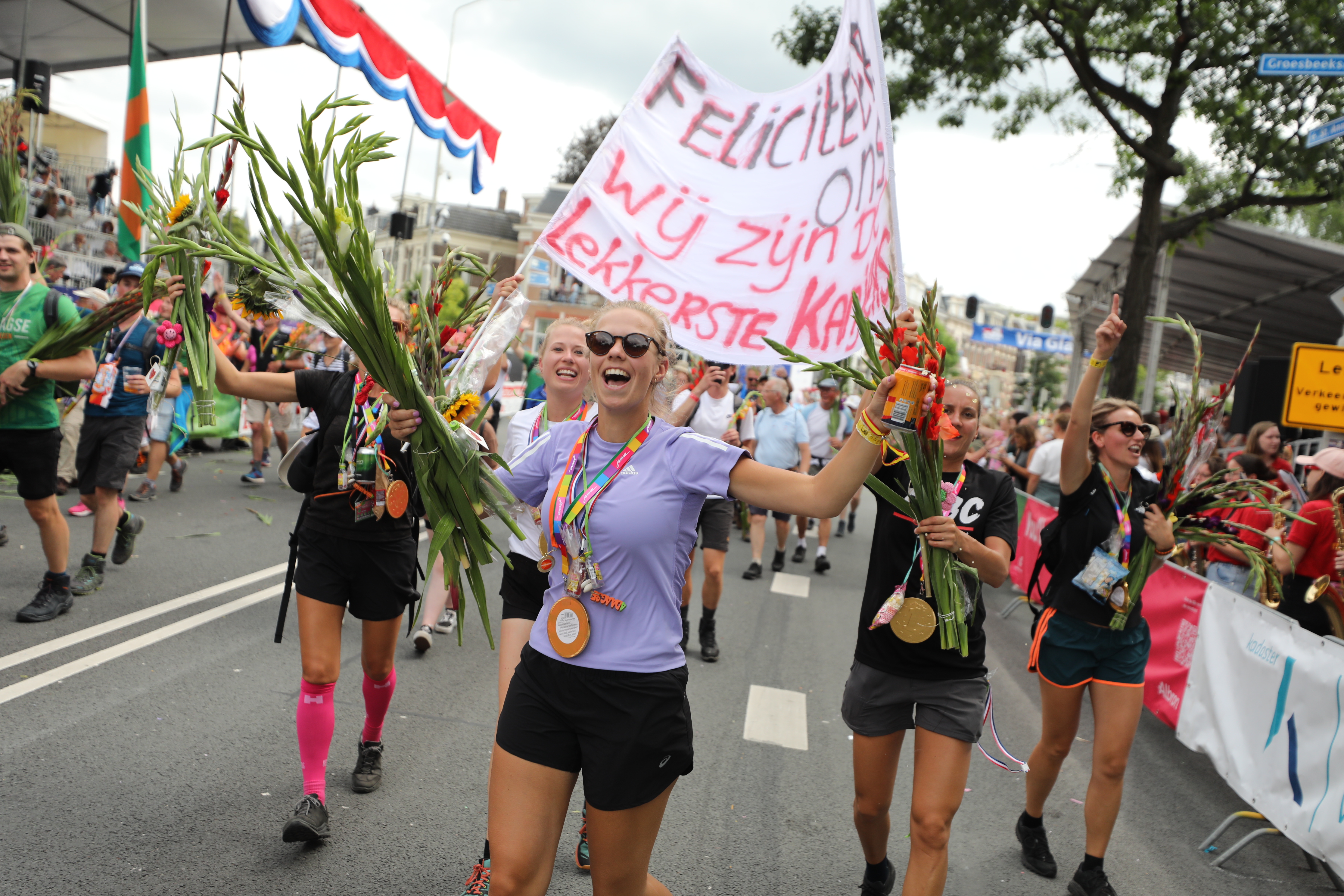 Juichende vrouwelijke deelnemers aan de Vierdaagse zijn net gefinished en dragen hun medaille en hebben gladiolen en een groot spanddoek vast.
