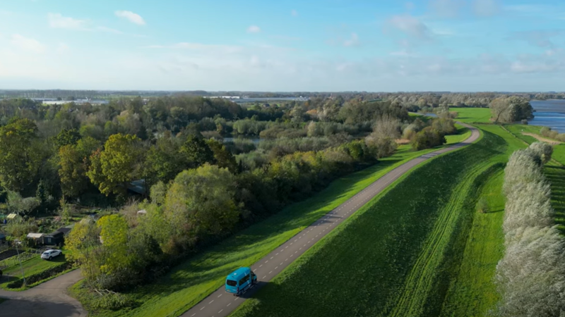 Filmstill van een bovenaanzicht van en weg op een dijk met daarop een groene Arriva-buurtbus. Filmstill van een bovenaanzicht van en weg op een dijk met daarop een groene Arriva-buurtbus.