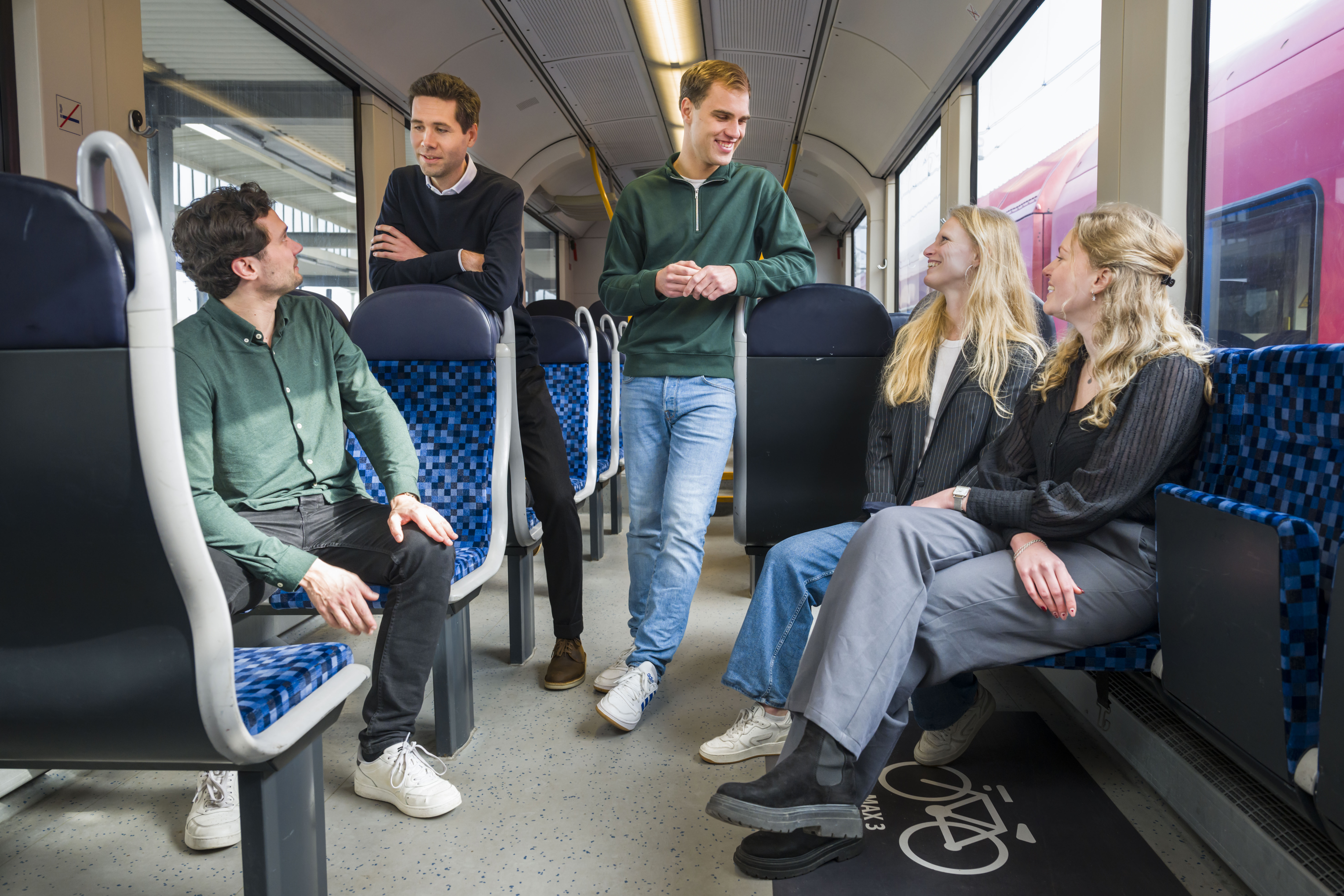 Vier personen in Arriva-trein, twee zittend, twee staand, in levendige discussie. Gekleed in smart casual, duidt op werkgerelateerde reis.